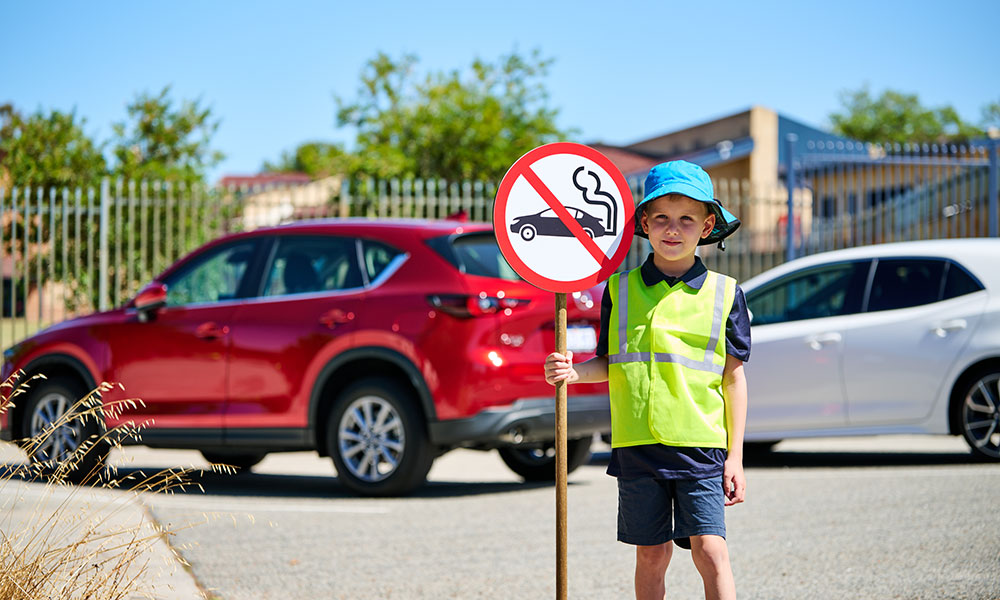 A young boy is standing in front of two parked cars, wearing a hi-vis vest and holding a sign of a car with exhaust coming from its tailpipe, indicating this is not permitted.