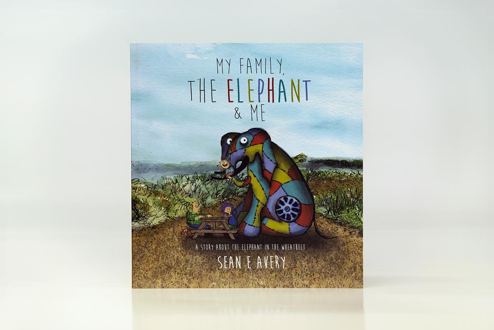 Front cover of 'My Family, the Elephant & Me' - A multicoloured elephant made of scrap metal sits on its hind legs while a child rides its tusks. Parents are seated on a picnic bench with their brown dog.