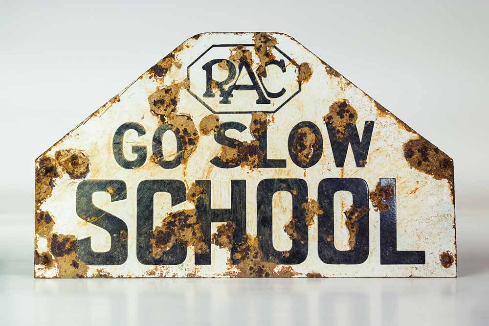 An old, rusted sign from the 1930s stating "Go slow; School." Above the text is the old RAC logo.