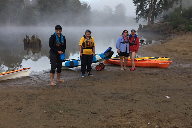 A small tourist group stand in front of kayaks on a river bank