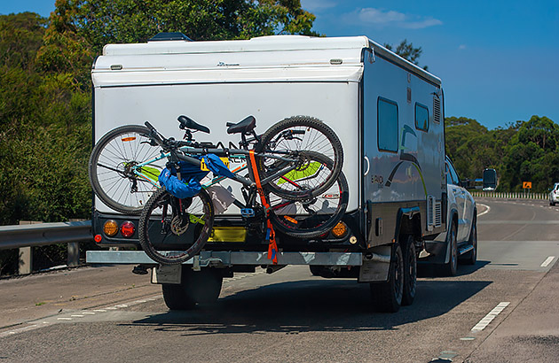 A caravan with a bike rack and bikes on the rear being towed on a country road