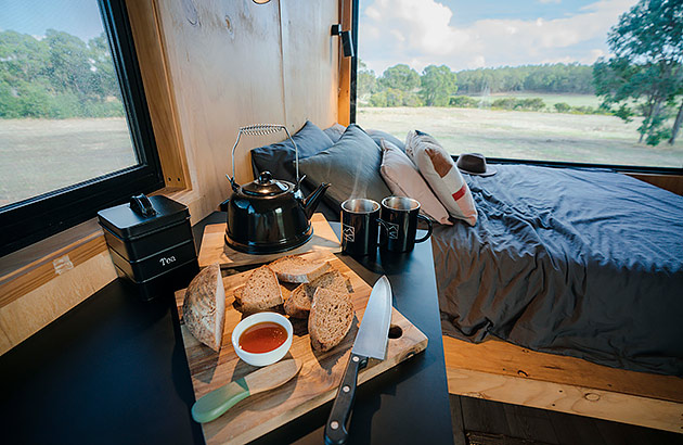 The interior of a tiny cabin showing the bed and view of a field thorough 2 windows