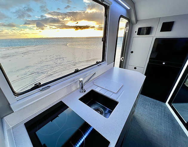 The kitchen of a Bruder caravan with a view out of the window to a beach