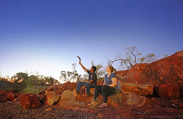  A traditional owner holding a boomerang is sitting next to a woman and pointing at the night sky