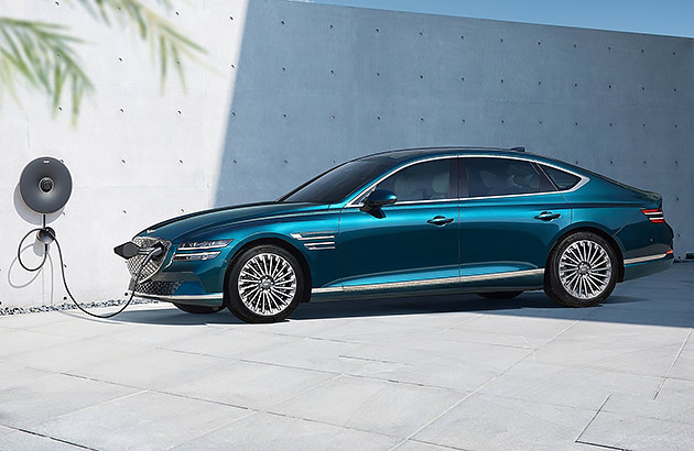 A blue Genesis G80 Electrified being charged with a white concrete wall in the background
