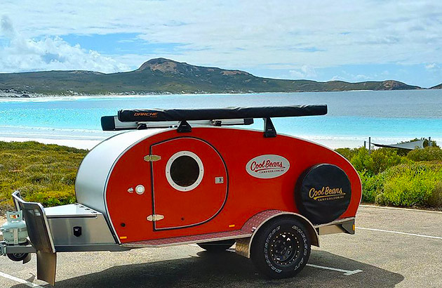 An orange Cool Beans brand teardrop camper parked near the beach