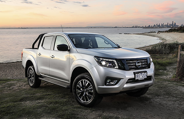 A white Nissan Navara parked by a shoreline with a city in the distance