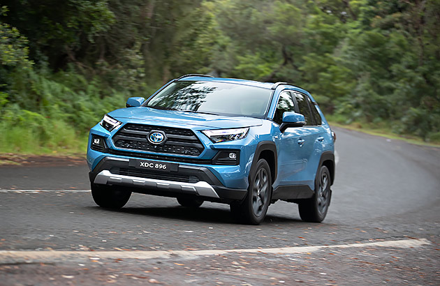 A light-blue Toyota RAV4 driving on a country road edged with green foliage