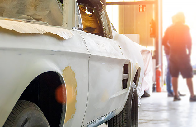 A stripped back car in the foreground of a workshop with two people standing in the doorway