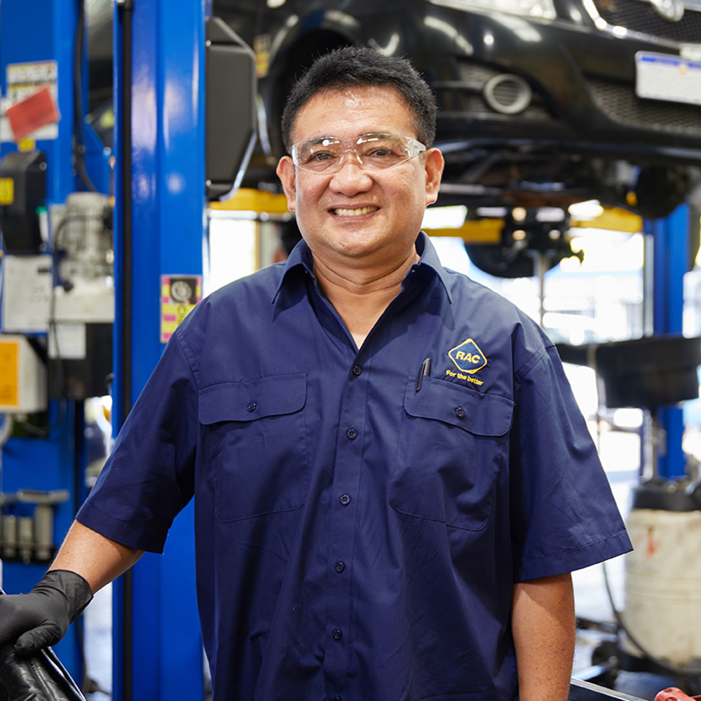 An RAC auto mechanic is wearing safety glasses, standing in front of a car smiling