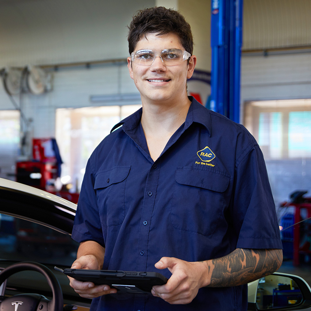 A young male RAC car mechanic is holding a tablet and standing in front of a Tesla