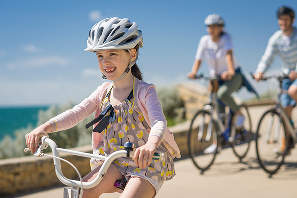 A young girl is smiling while riding her pushbike, and two adults are riding their bikes behind her