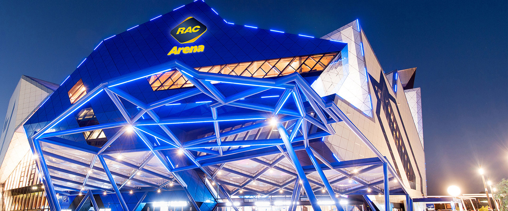 The RAC Arena is lit up at night, showcasing the blue structure against the Perth skyline.