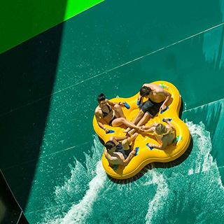 Four people sitting on a bright yellow tube, racing down a green water slide at Adventure World