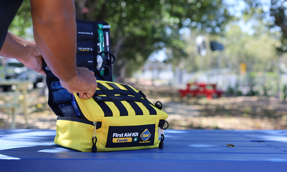 A person is opening up a bright yellow RAC Family First Aid Kit on a blue park bench