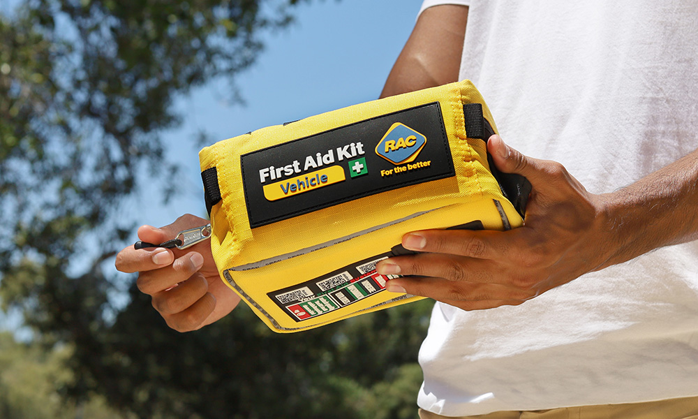 A person is holding a bright yellow RAC Vehicle First Aid Kit outside