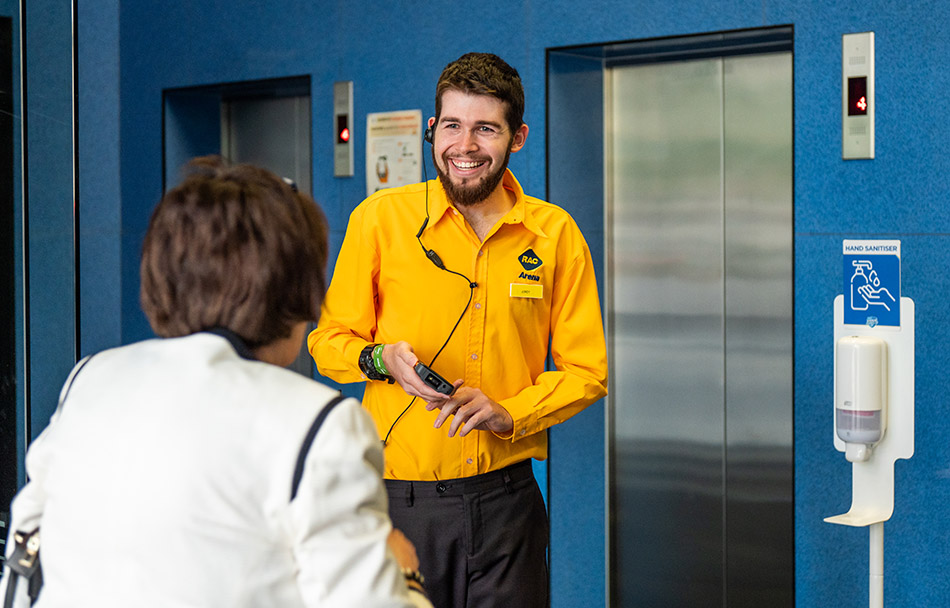 An RAC Arena colleague is smiling and greeting a visitor in front of the lifts