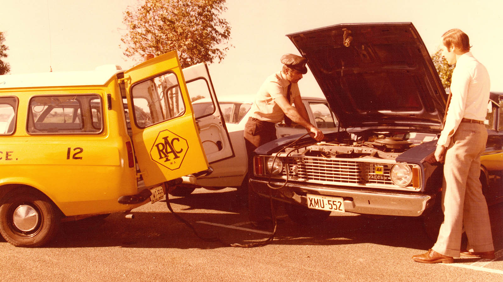 A historical photo of an RAC Mobile Technician looking under the bonnet of a parked car, with a man watching on from the side 
