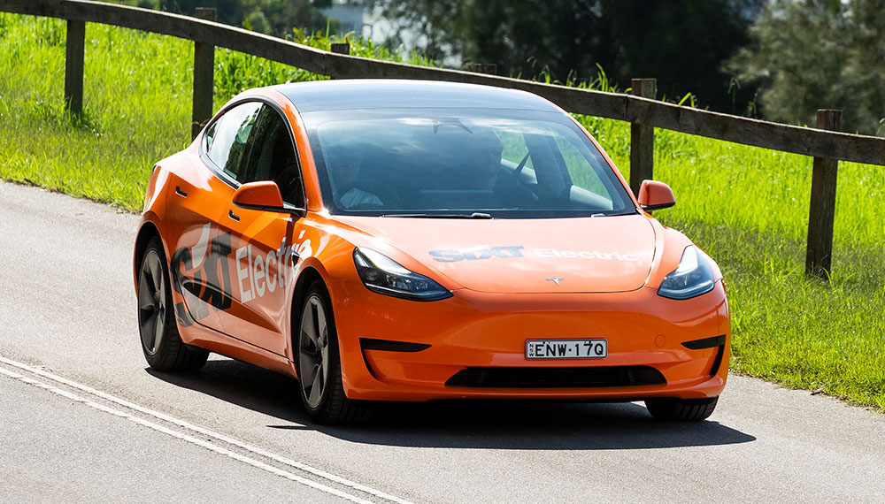 A bright orange electric car with the word SIXT written across the doors is driving next to a green paddock