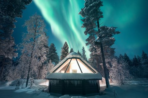 A hut with big panoramic ceiling windows standing in the middle of a snowy landscape with glowing Northern Lights in the backhround