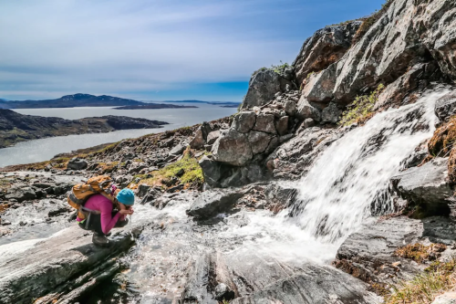  A hiker drinks from a mountain stream near a waterfall with a scenic backdrop of rocky terrain and distant lakes.