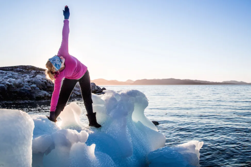  A person stands on an ice formation by the sea, stretching with one arm raised under a clear sky.