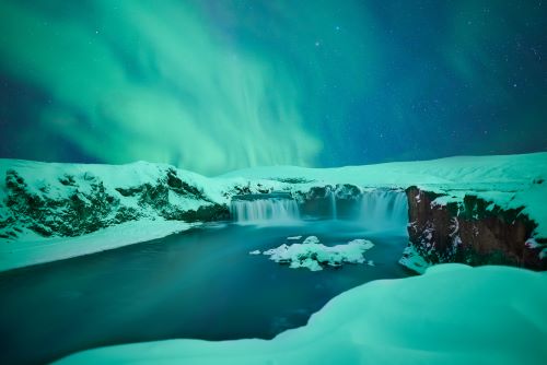 A snowy landscape features a waterfall and a river under a sky illuminated by the Northern Lights.