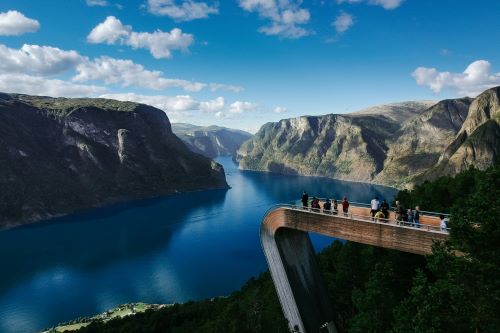 People stand on a viewing platform overlooking a stunning fjord surrounded by mountains.