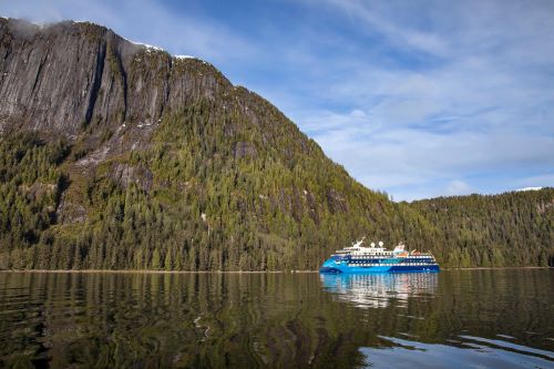 The American Queen Vessel looking small next to the steep mountain cliffs covered with pine trees and snowy tips