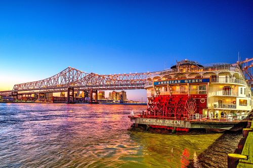 The American Queen vessel with its red paddle wheel docked in New Orleans at dusk with a long truss bridge in the background 