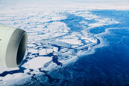 An airplane engine in the foreground with a vast, frozen landscape of broken ice and snow-covered terrain below.