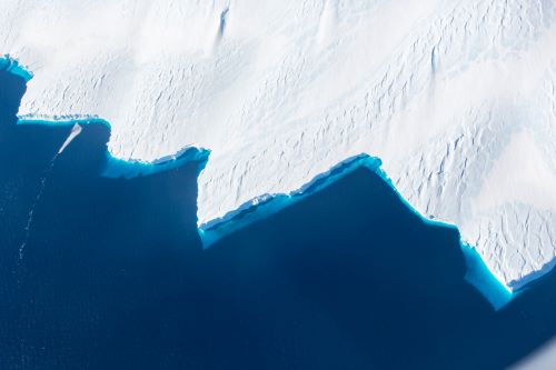 A dramatic aerial view of a snow-covered coastline with jagged edges meeting a deep blue ocean.