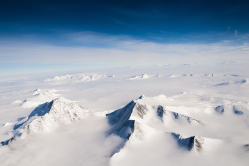 Snow-covered peaks breaking through a cloud cover above the vast Antartica landscape. 