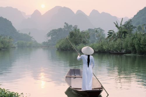 A woman in a plain white dress and typical Vietnamese hat is rowing down the river in a small wooden boat 