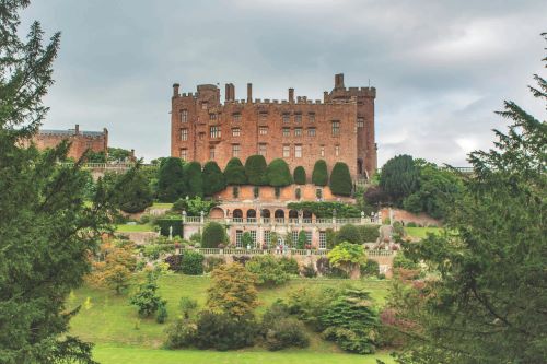 A large, historic brick castle surrounded by manicured gardens and lush greenery.