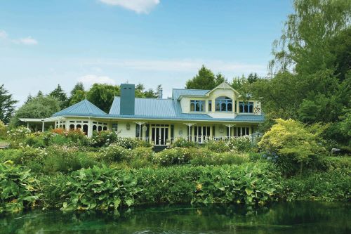 A beautiful homestead with light-blue roof close to a pond and surrounded by lush greenery and flowers. 