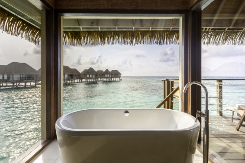 A free standing bathtub right by the windows overlooking the water bungalows and ocean in the background