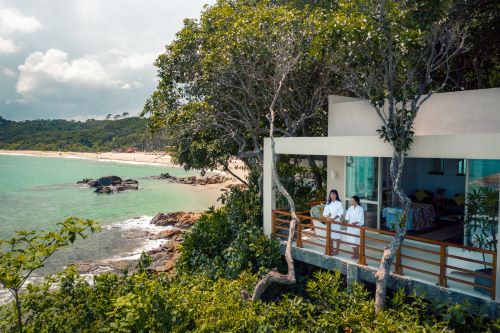 A couple of visitors standing on the balcony of their accommodation surrounded by lush nature and are looking out at the ocean 