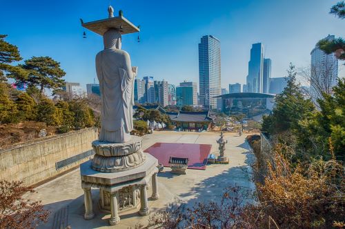 A traditional Korean Buddhist temple complex with a tall Buddha statue facing the skyline of Seoul on a sunny day