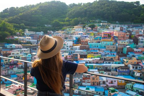 A woman with a hat facing her back t the camera is standing at a lookout overlooking the many colourful houses of Busan Gamcheon Culture Village