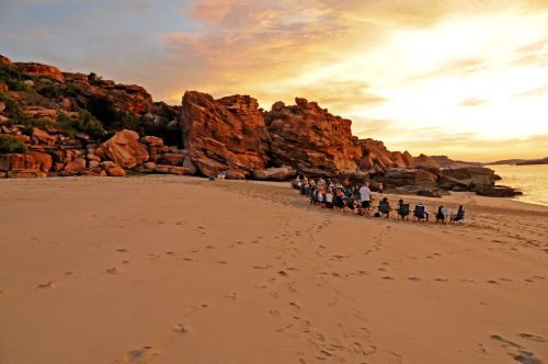 A group of passengers sitting on camping chairs at the beach watching the sunset surrounded by spectacular red cliffs typical for the Kimberley region