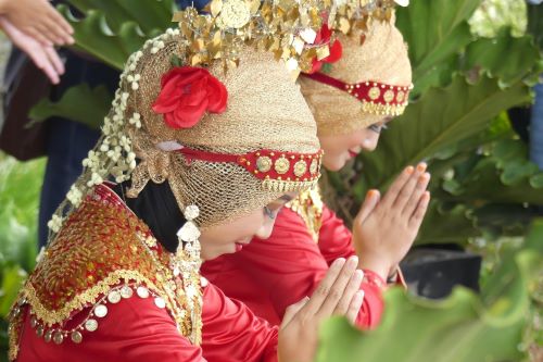 Two women from Belitung folding their hands for prayer with closed eyes are wearing traditional red and gold costumes with a golden headscarf decorated with beads, gold headpiece and a red flower.