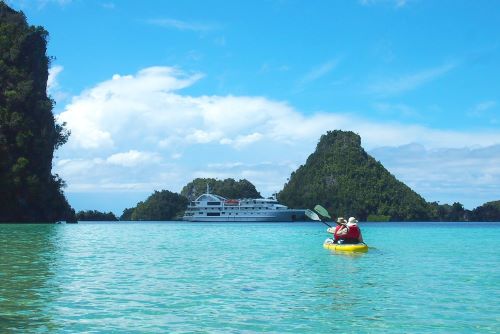 A couple in a kayak paddling through turquoise water towards the Coral Expeditions vessel which is at bay between some high rising islands in the background