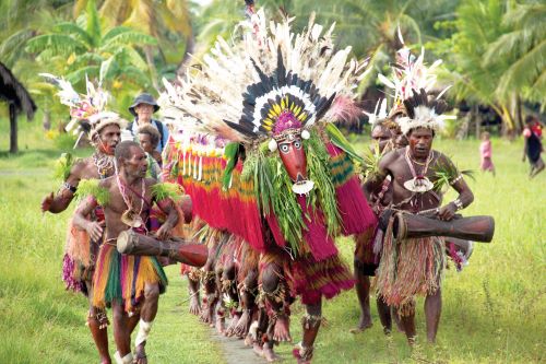 Indigenous people from Papua New Guinea walking down in three rows playing the drums and singing while some are dancing while  wearing big colourful masks with lots of feathers