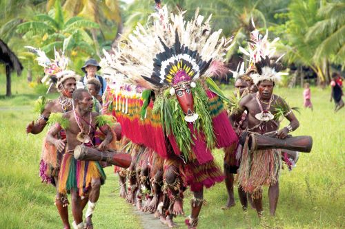 Indigenous people from Papua New Guinea walking down in three rows playing the drums and singing while some are dancing while wearing big colourful masks with lots of feathers