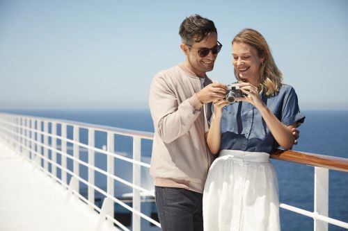 Couple standing aboard Cunard vessel looking at photos they have taken with their camera and smiling