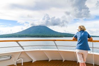 A woman standing at the back of the ship with her back facing the camera overlooking the scenery in front of her