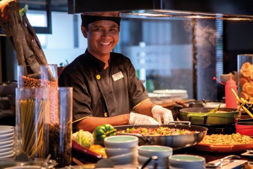 A smiling chef prepares colorful dishes at a buffet station with vibrant ingredients on display.