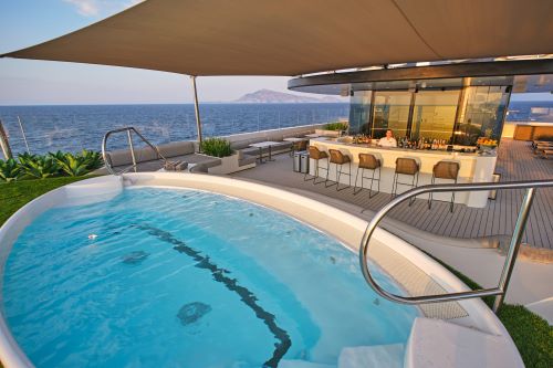 A luxurious rooftop pool and bar area on a cruise ship, featuring a round spa pool with ocean views, a shaded seating area, and a modern bar with high stools.
