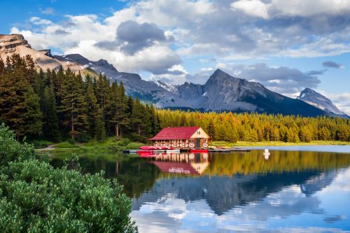 A wooden hut with a red roof by a serene lake surrounded by mountains and forest